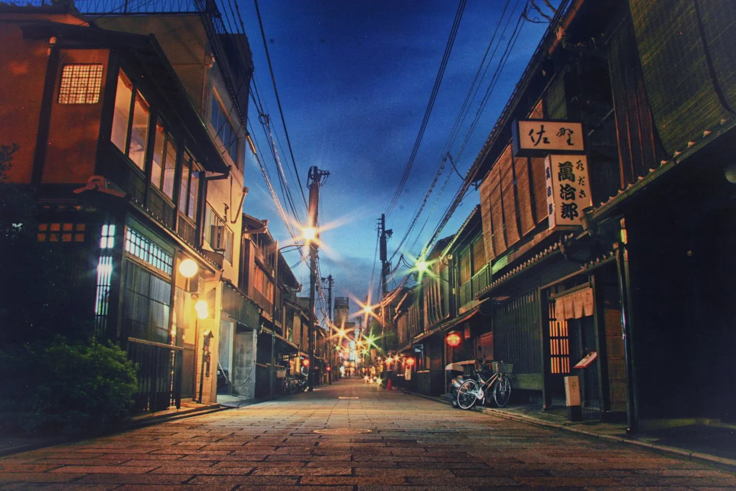 夜明け前の石畳の通り。町家の軒先に灯りが残っている。